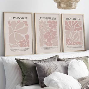 May include: Three framed art prints with floral designs in shades of pink and beige. Each print features a different Bible verse: Romans 8:28, Jeremiah 29:11, and Proverbs 3:5. The prints are displayed above a bed with decorative pillows.
