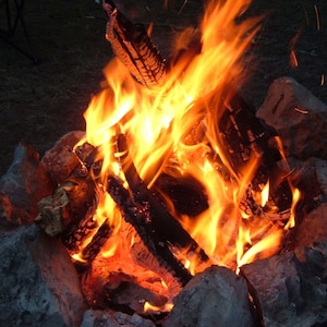 May include: A close-up of a campfire burning brightly with orange and yellow flames. The fire is surrounded by rocks and there are some logs in the fire.