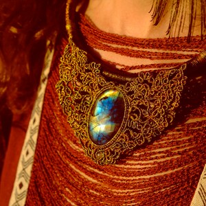 May include: A gold necklace with a large, iridescent blue gemstone pendant. The necklace is intricate and detailed, with a delicate, lacy design. The pendant is oval-shaped and has a shimmering, multi-colored effect.
