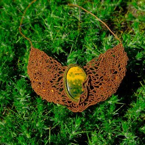 Può includere: Una collana in macramè marrone con un grande ciondolo in agata verde e giallo. La collana è appoggiata su un letto di erba verde.