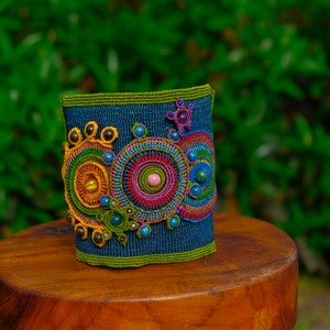 May include: A colorful beaded bracelet with a blue background and a circular pattern of beads in shades of yellow, orange, red, pink, green, and blue.