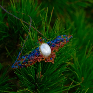 May include: A colorful macrame necklace with a white iridescent stone pendant. The necklace is made with a variety of colors, including red, orange, yellow, green, blue, and purple.