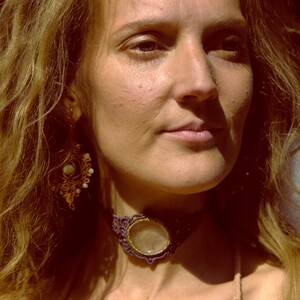 Puede incluir: Una mujer con el pelo largo, ondulado y castaño claro lleva un collar de cuero marrón con un gran colgante redondo de piedra preciosa transparente. También lleva un par de pendientes de oro con un diseño floral.