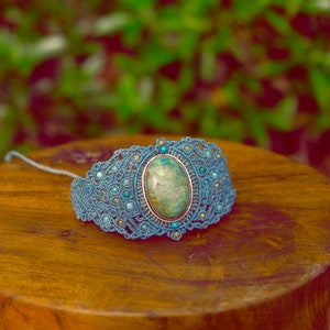 May include: A blue macrame bracelet with a green and brown oval gemstone in the centre. The bracelet is decorated with small blue beads.