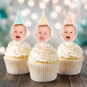 May include: Three cupcakes with white frosting and small white sprinkles, each topped with a paper cutout of a baby's face wearing a party hat. The hats are light blue, yellow, and light pink. The babies have different facial expressions.