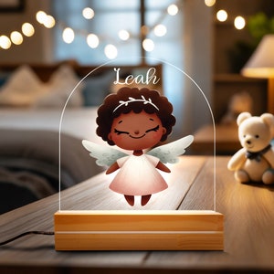 May include: A personalized night light featuring a cartoon angel with the name "Leah" in script. The angel has dark skin, a white dress, and wings. The light sits on a wooden base and is on a wooden table with toys in the background.