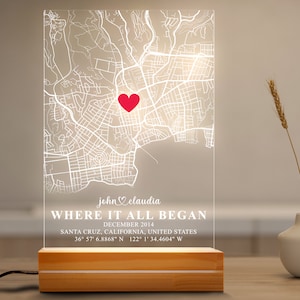 May include: A clear acrylic light-up map with a red heart marking a location. The text below reads "john claudia WHERE IT ALL BEGAN DECEMBER 2014 SANTA CRUZ, CALIFORNIA, UNITED STATES 36° 57' 6.8868" N 122° 1' 34.4604" W". The map is on a wooden base.