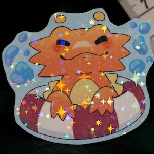 May include: A holographic sticker featuring an orange lizard-like creature with a smiling expression, floating in a red and white striped inner tube surrounded by blue bubbles. A measuring tape is visible, showing a width of approximately 6.5 cm.