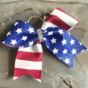 May include: A patriotic bow with a silver key ring. The bow features blue glitter fabric with white stars and red and white striped glitter fabric. A sparkling rhinestone embellishment is at the centre of the bow.