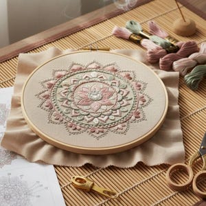 May include: A circular embroidery hoop holding a finished mandala design in pink, green, and beige thread. The embroidery is on beige fabric. Spools of thread and scissors are visible, indicating a craft project or a DIY kit.
