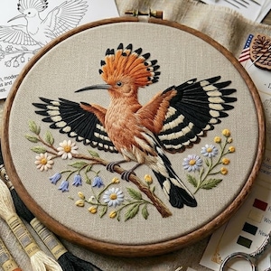 May include: An embroidered Hoopoe bird with open wings, perched on a branch with flowers, displayed in a wooden hoop. The bird's feathers are orange, black, and white, set against a beige fabric. Embroidery threads and other supplies are also visible.