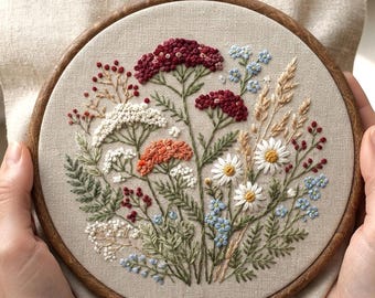 May include: A circular wooden embroidery hoop containing a beige fabric panel with a detailed hand-stitched floral arrangement. The design features clusters of red, orange, white, and light blue flowers, green foliage, and tan wheat stalks.