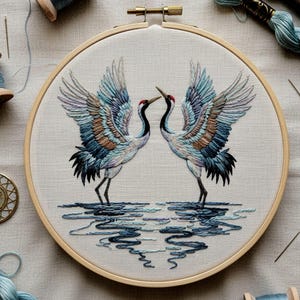 May include: Embroidered artwork in a wooden hoop features two cranes facing each other. The birds have blue, brown, and white feathers, with red crowns. The water reflection is in shades of blue. The background is a light beige fabric.