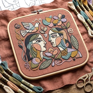 May include: A square, hand-stitched embroidery hoop featuring two stylized faces, a bird, and floral elements in muted tones of peach, teal, and gold. The artwork is framed in a natural wood hoop, with embroidery floss and scissors nearby.