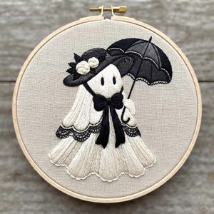 Könnte beinhalten: Ein runder hölzerner Stickrahmen mit einem Stoffstück, das eine handgestickte Geisterfigur zeigt. Der Geist trägt einen schwarzen Hut mit weißen Blumenakzenten, eine schwarze Fliege und hält einen schwarzen Regenschirm in der Hand.