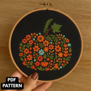 以下が含まれることがあります： 木製のフレームに刺繍されたカボチャのデザイン。カボチャは、黒い背景にオレンジ、青、緑の花と葉で満たされています。「PDF PATTERN」の文字が左下にあります。フレームは手で持たれています。