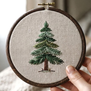 Puede incluir: Un bastidor de bordado circular con un marco de madera oscura que contiene una tela de color beige claro. Centrado en la tela hay un árbol de hoja perenne bordado a mano con varios tonos de hilo verde para las agujas y marrón para el tronco.