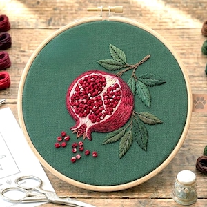 Peut inclure: Broderie artisanale représentant une grenade, avec des détails en rouge et rose. Le fruit est mis en valeur sur un fond vert foncé, encadré dans un cercle à broder en bois. Des feuilles vertes et des graines éparpillées complètent le motif, pour une œuvre texturée.