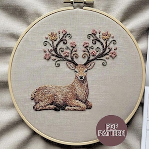 Puede incluir: Bordado de un ciervo con cuernos florales, sobre tela beige, en un aro de madera. El ciervo es marrón, con flores rosas y ramas marrones que forman los cuernos. Una etiqueta circular dice "PDF PATTERN".
