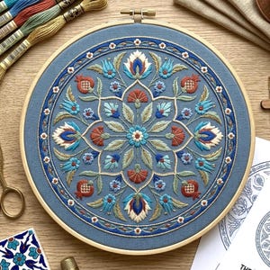 May include: A circular embroidery hoop containing blue fabric with a symmetrical floral and pomegranate pattern stitched in shades of blue, red, white, and green. The design features a central flower surrounded by intricate petals and leaves.