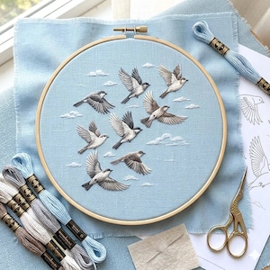 May include: An embroidery hoop with a light blue fabric featuring a flock of embroidered birds in flight. The birds are gray and white, with detailed wing patterns. Embroidery floss in various shades of blue and gray are visible.