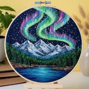 May include: Embroidered hoop art featuring a nighttime landscape with a vibrant aurora borealis. The design includes snow-capped mountains, a dark blue lake, and a forest of green trees. The sky is a deep blue with white stars.