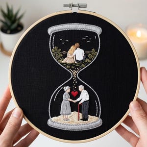 May include: Embroidered artwork in a wooden hoop. The design features an hourglass with a young couple at the top and an elderly couple at the bottom, connected by a stream of dots. A red heart is between the elderly couple.