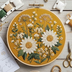 May include: A handcrafted embroidery hoop featuring a floral design with white daisies, bell-shaped flowers, and other greenery on a mustard yellow fabric. The wooden hoop is surrounded by spools of thread and a stitch guide.