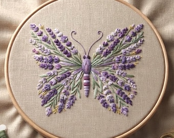 Puede incluir: Un bastidor de bordado circular de madera que contiene un panel de tela beige. La tela presenta un diseño de mariposa bordado a mano compuesto por ramas de lavanda moradas y hojas verdes, con un cuerpo central morado y dos antenas finas.