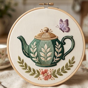 May include: Embroidered tea-themed hoop art featuring a green teapot with leaf and floral details, a butterfly, and a floral garland. The embroidery is set against a cream-colored fabric within a wooden hoop, creating a charming, handcrafted aesthetic.