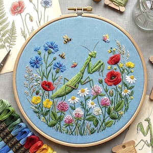 May include: A circular wooden embroidery hoop containing a light blue fabric panel with a detailed hand-stitched design of a green praying mantis surrounded by colorful wildflowers, including red poppies, blue cornflowers, and white clover.