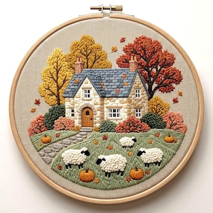 May include: A circular embroidery hoop containing a fabric panel with a stitched scene of a stone cottage, autumn trees in yellow and orange, and four white sheep grazing on a green lawn with scattered orange pumpkins and fallen leaves.