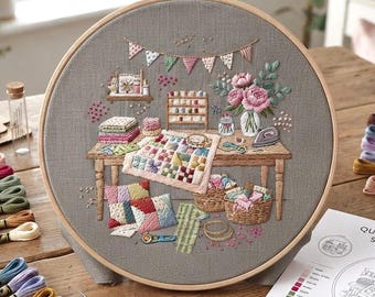 May include: A circular wooden embroidery hoop containing gray fabric with a detailed needlepoint scene of a craft room. The design features a wooden table, a patchwork quilt, a vase of pink flowers, thread spools, baskets, and colorful bunting.