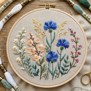 May include: A circular wooden embroidery hoop containing a linen fabric piece with a hand-stitched floral design. The embroidery features blue cornflowers, peach-coloured blossoms, small white flowers, and pink sprigs, surrounded by green stems.