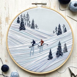 May include: Embroidered winter scene in a wooden hoop. The artwork depicts two skiers on a snowy slope, surrounded by dark green pine trees. The background is a light blue fabric, and the hoop is made of wood.