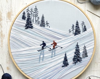 Könnte beinhalten: Gestickte Winterszene in einem Holzreifen. Das Kunstwerk zeigt zwei Skifahrer auf einer schneebedeckten Piste, umgeben von dunkelgrünen Kiefern. Der Hintergrund ist ein hellblauer Stoff, und der Reifen ist aus Holz.