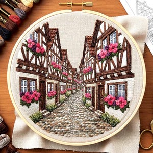 May include: Embroidered artwork in a wooden hoop depicts a quaint European street scene. The buildings feature brown timber framing, white walls, and red-tiled roofs. Pink flowers adorn the window boxes, and a cobblestone street leads into the distance.
