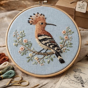 May include: An embroidered hoop art piece showing a hoopoe bird on a branch with flowers. The bird has brown, black, and white markings, and a crest. The background is light blue fabric, and the hoop is made of wood. The embroidery is detailed.