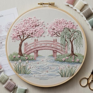 May include: Embroidered artwork in a wooden hoop shows a pink bridge over water, with cherry blossom trees. The scene includes green grass, water ripples, and pink flowers. Embroidery floss and a pattern are visible.