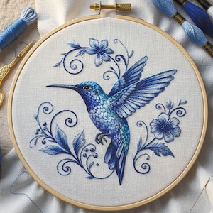 May include: A detailed embroidery of a hummingbird in flight, showcasing shades of blue and teal. The bird is surrounded by floral and leaf designs, all within a wooden embroidery hoop. Embroidery floss and gold scissors are visible.