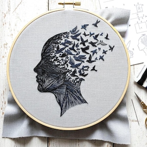 Op de afbeelding: Geborduurd kunstwerk in een houten ring. Het ontwerp toont een profiel van een gezicht, waarbij het hoofd overgaat in een zwerm donkerblauwe vogels. De achtergrond is lichtgrijze stof.