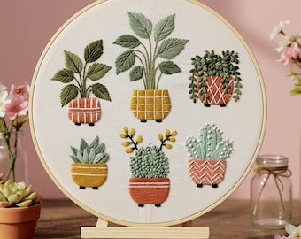 May include: Embroidered hoop art featuring six potted plants. The plants are in various styles, with green leaves and colorful pots. The embroidery is on a white fabric, displayed on a small wooden easel.
