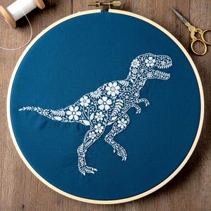 May include: A blue fabric embroidery hoop featuring a white floral T-Rex design. The embroidery is set in a wooden hoop, with a spool of white thread and gold scissors in the background. The design is a unique blend of nature and prehistoric themes.