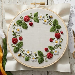 Könnte beinhalten: Gestickter Erdbeerkranz auf weißem Stoff in einem Holzreifen. Das Design umfasst rote Erdbeeren, grüne Blätter, weiße Blüten, einen Marienkäfer und eine Biene. Die detaillierte Stickerei zeigt ein handgefertigtes Kunsthandwerk.