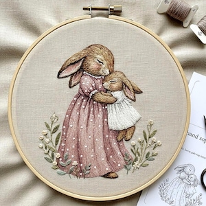 Può includere: Ricamo su telaio raffigurante due coniglietti in un tenero abbraccio. Il coniglietto più grande indossa un vestito rosa a pois bianchi, mentre il coniglietto più piccolo indossa un vestito bianco. Il disegno è circondato da verde e fiori ricamati.