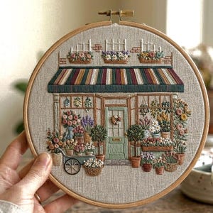 Può includere: Scena ricamata di un negozio di fiori in un telaio di legno. Il design presenta una porta verde, una tenda a strisce e vari fiori in vaso. La tavolozza dei colori include verdi, marroni, rossi e crema, creando un'opera d'arte dettagliata.