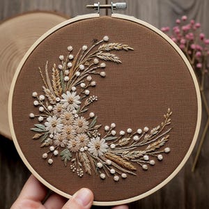 Op de afbeelding: Een bruine borduurring met een bloemenontwerp. De borduursel toont een halvemaanvormige opstelling van witte madeliefjes, tarwe en kleine witte kralen. De stof is effen bruin en de ring is van hout.