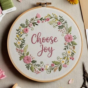 Op de afbeelding: Een borduurring met de tekst "Choose Joy" geborduurd in roze draad. Het ontwerp toont een krans van roze rozen, witte bloemen en groene bladeren. De ring is gemaakt van lichtgekleurd hout.