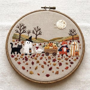 May include: A circular wooden embroidery hoop containing a fabric panel with a hand-stitched autumn scene. It features a black cat, a ghost in a witch hat, a white goose, a wooden cart filled with orange pumpkins, a fox, and a second ghost.