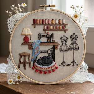Könnte beinhalten: Gestickter Stickrahmen mit einer Nähzimmer-Szene. Eine schwarze Katze liegt auf einem Kissen vor einer Nähmaschine. Die Szene umfasst eine kleine Lampe, einen Hocker, Schneiderpuppen und Garnrollen. Der Hintergrund ist ein neutraler Leinenstoff.